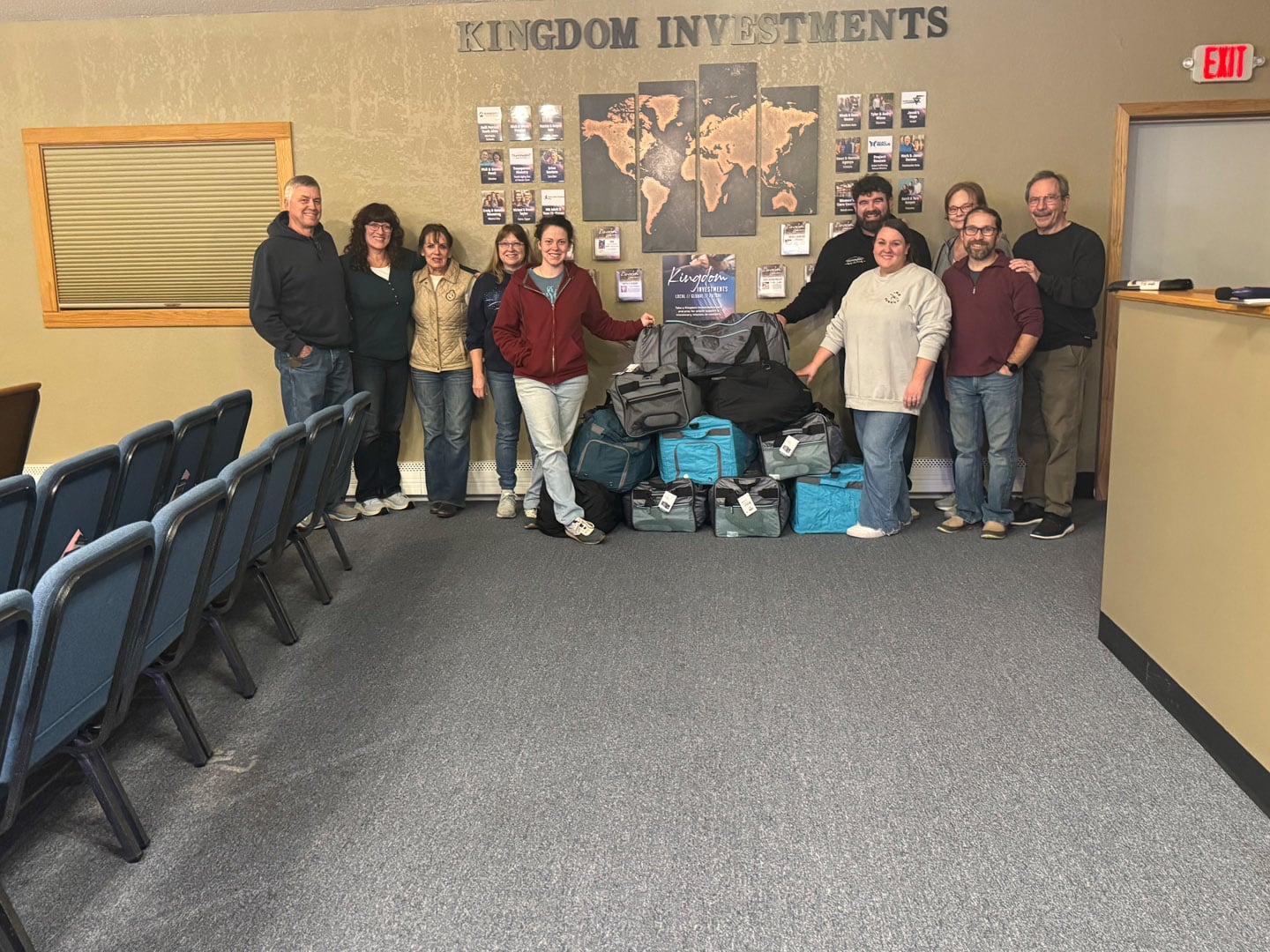 A group of ten people stands in a room next to a wall display labeled "Kingdom Investments," posing with several bags and boxes stacked in front of them. Rows of blue chairs are visible on the left.