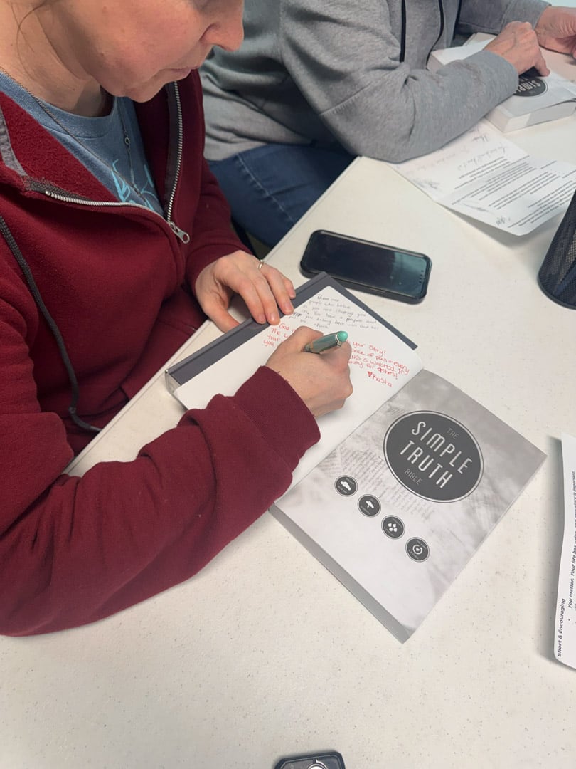 A person in a red jacket writes notes in red ink inside a book titled "Simple Truth" on a table with a smartphone and papers nearby.