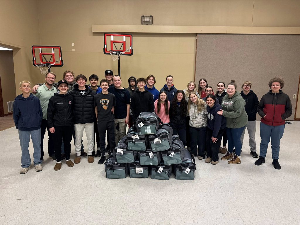 A group of about 25 young people and adults stand indoors behind a pyramid-shaped stack of duffel bags. Two basketball hoops are visible on the wall behind them. Everyone is smiling, and the room has a gymnasium appearance.