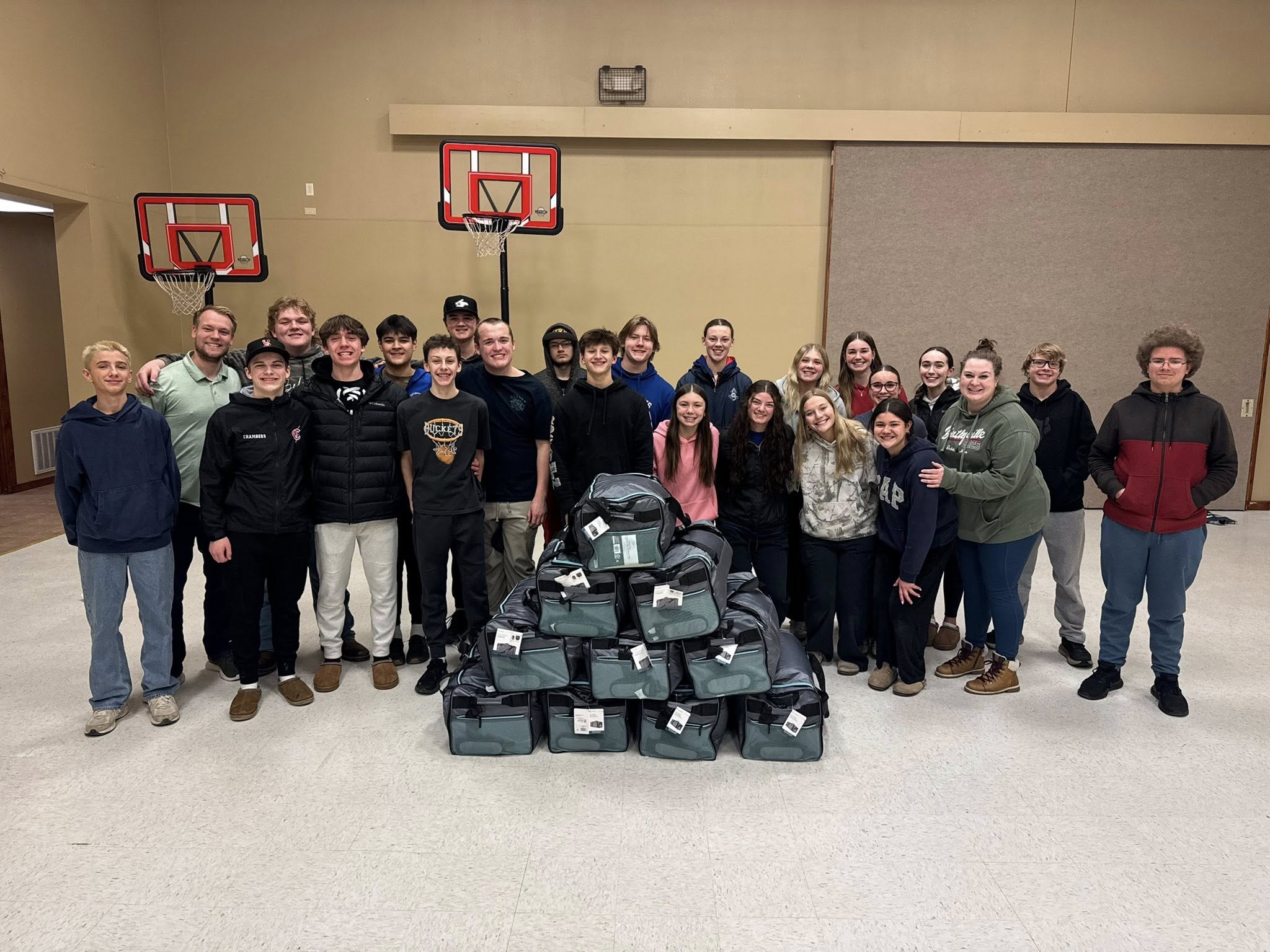 A group of about 25 young people and adults stand indoors behind a pyramid-shaped stack of duffel bags. Two basketball hoops are visible on the wall behind them. Everyone is smiling, and the room has a gymnasium appearance.