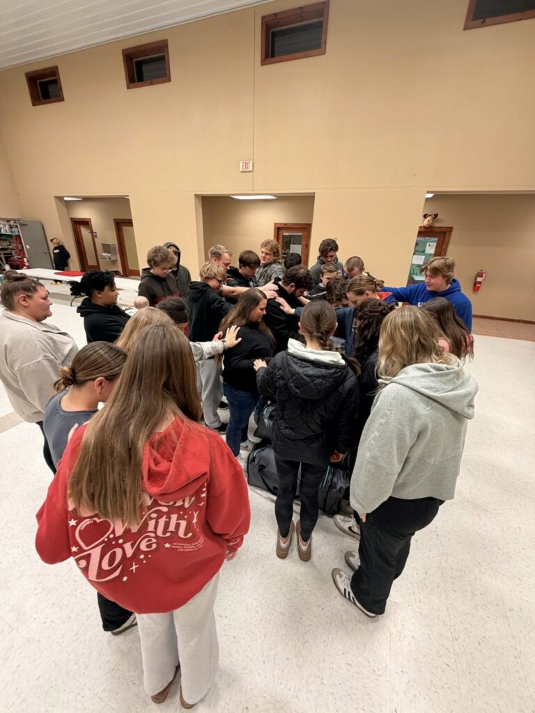A group of people stand closely together in a circle indoors, some with heads bowed and arms extended toward the center, in what appears to be a moment of prayer or support.