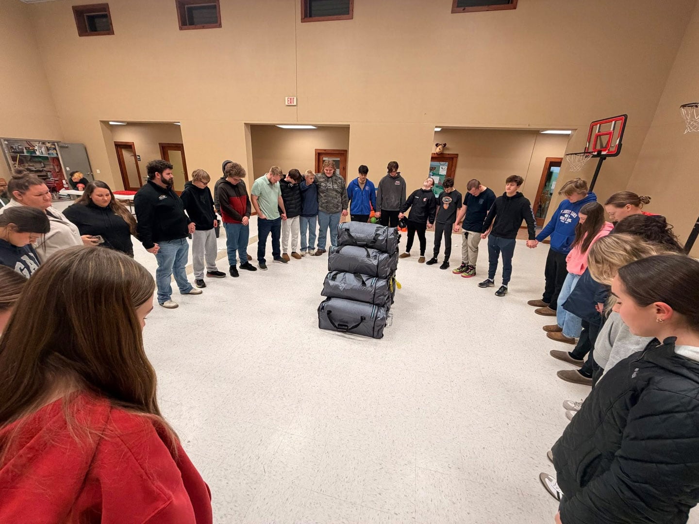 A group of young people stand in a circle holding hands, surrounding a stack of gray bags in the center of a large indoor room. They appear to be participating in a group activity or moment of reflection.