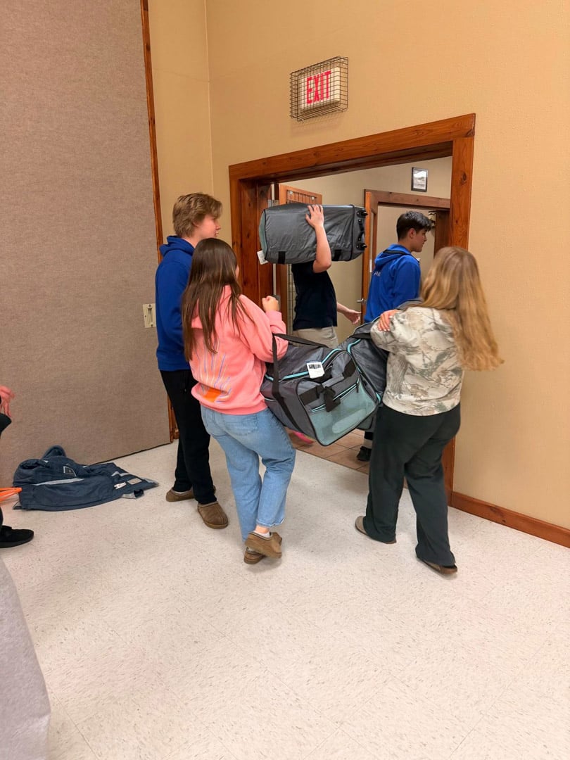 Four teenagers are carrying sleeping bags and duffel bags through a doorway in a building, heading outside. They are dressed casually, and the scene suggests they are preparing for a trip or event.