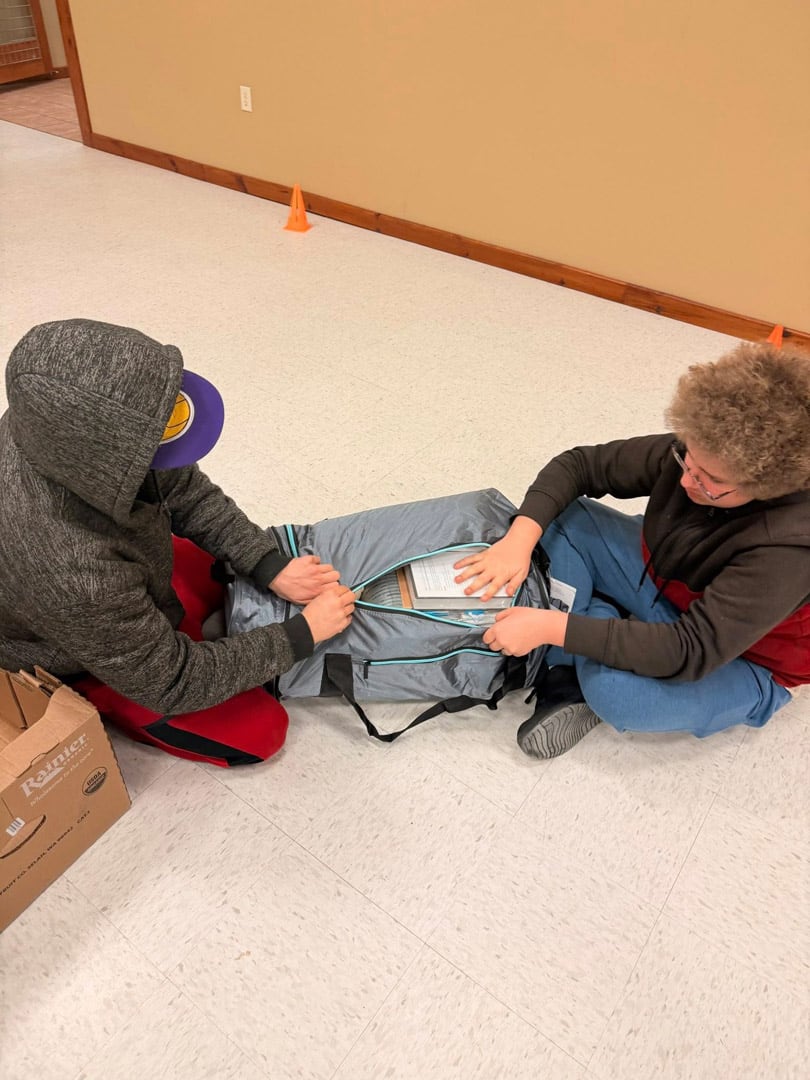 Two kids sit on the floor, packing or unpacking a gray bag together. One wears a gray hoodie and cap, the other wears glasses and a red and black jacket. There’s a cardboard box and orange cone nearby.