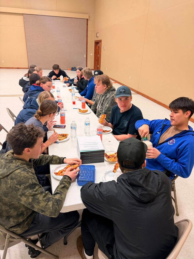 A group of teenagers sits around a long table in a large room, eating pizza and drinking bottled water. They are engaged in conversation and appear to be enjoying a casual meal together.
