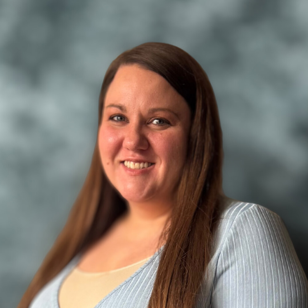 A woman with long brown hair smiles at the camera. She is wearing a light blue top and stands in front of a blurred gray-blue background.