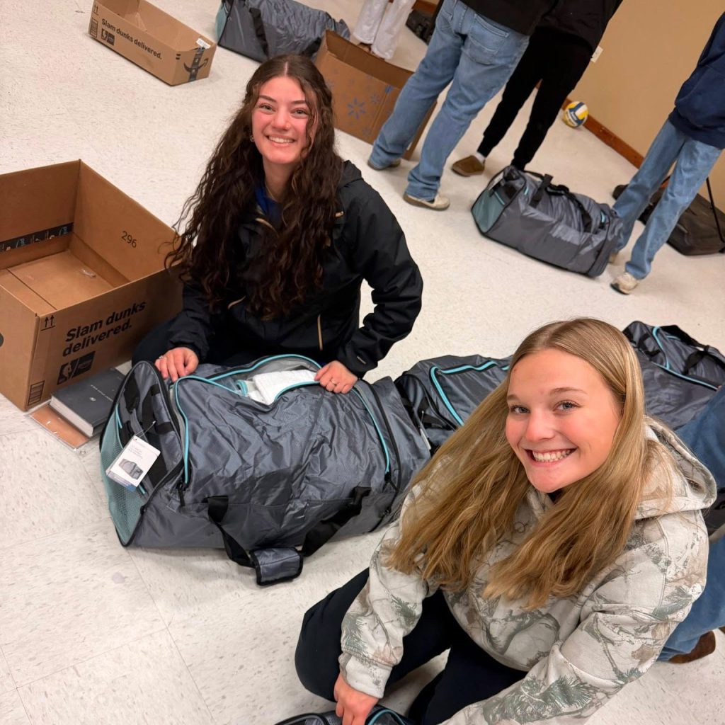 Two young women smile at the camera while kneeling on the floor, packing large gray duffel bags. Open cardboard boxes and more bags are scattered nearby. Other people stand in the background.