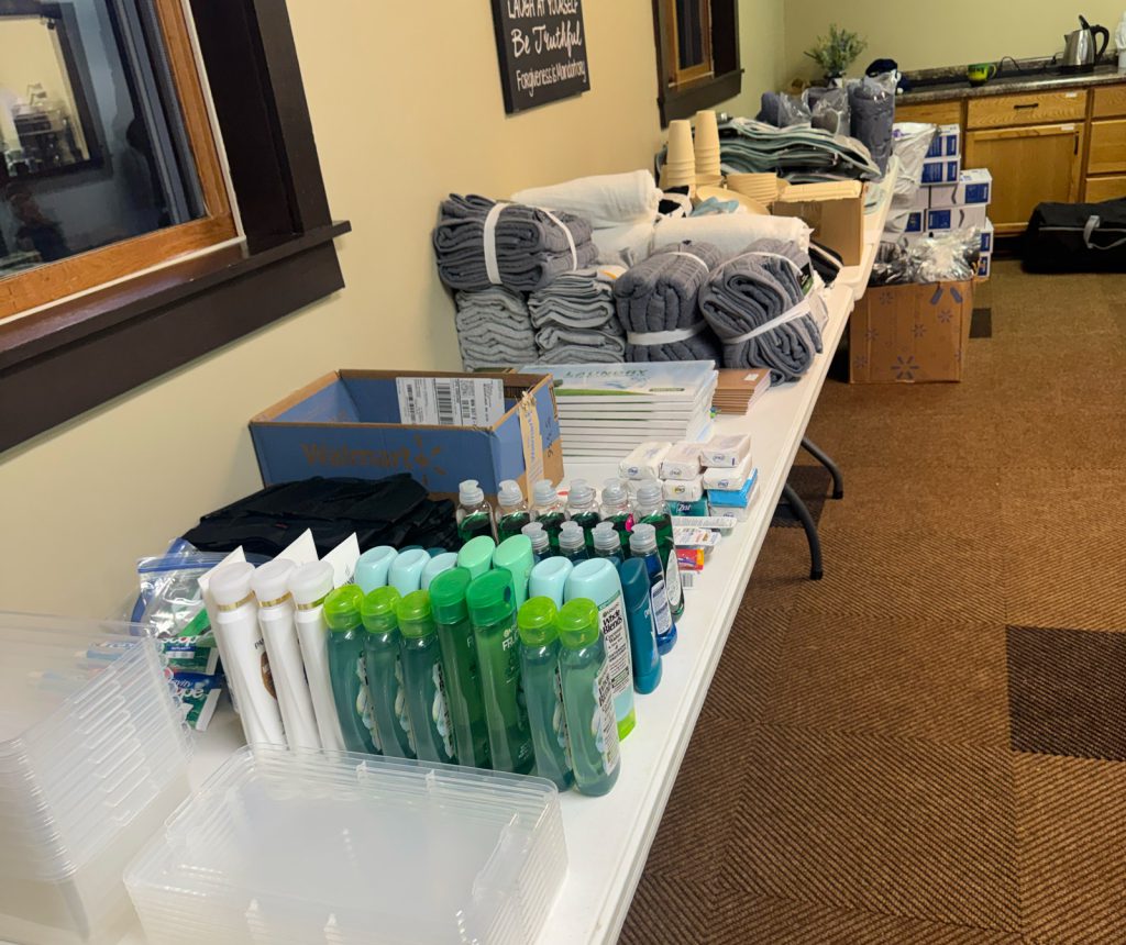 A long table displays neatly arranged toiletries, bottled liquids, rolled towels, stacked washcloths, cups, and other personal care supplies in a room with tan walls and a carpeted floor.