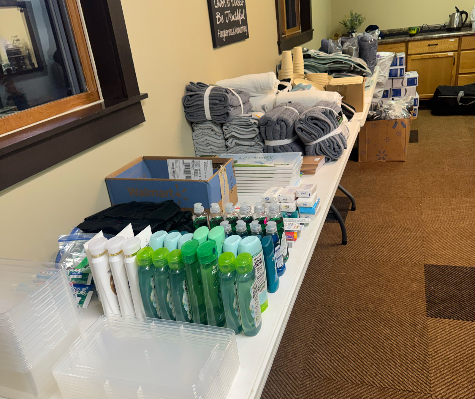 A long table displays neatly arranged toiletries, bottled liquids, rolled towels, stacked washcloths, cups, and other personal care supplies in a room with tan walls and a carpeted floor.