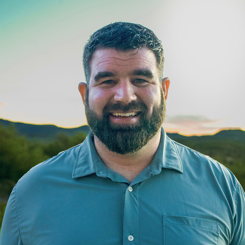 A man with short dark hair and a beard, wearing a light blue button-up shirt, smiles at the camera outdoors with hills and a bright sky in the background.