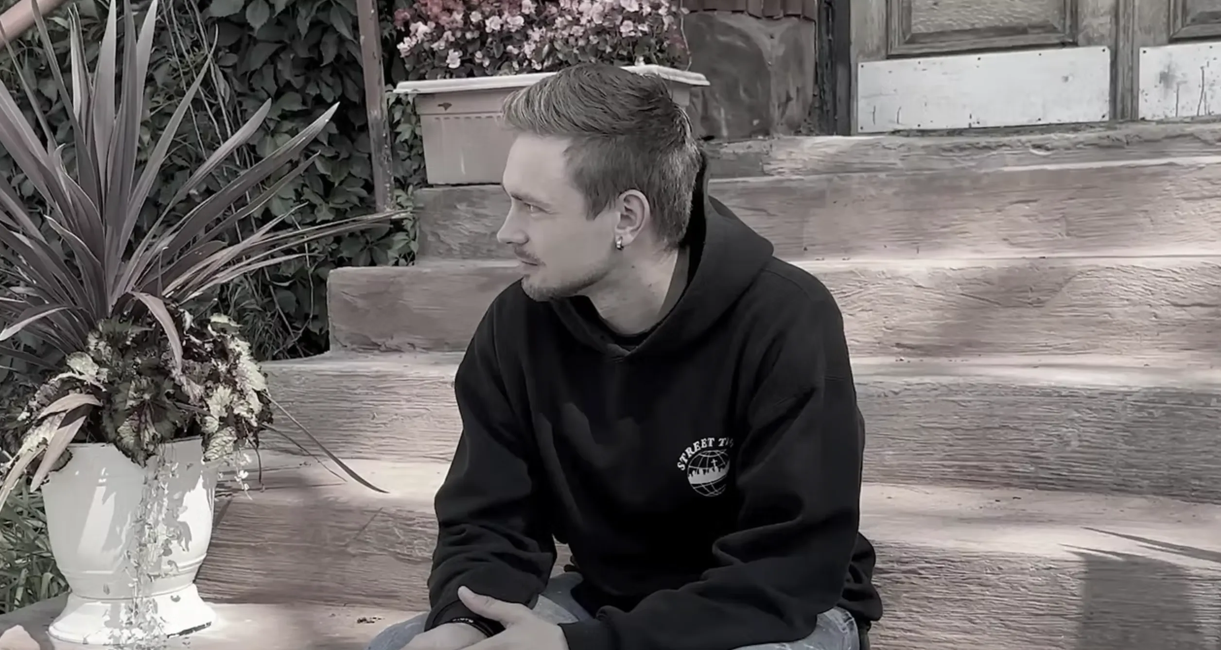 A man in a black hoodie sits on outdoor wooden steps, looking to the side. There are potted plants and flowers beside him and behind him, with a weathered door in the background.