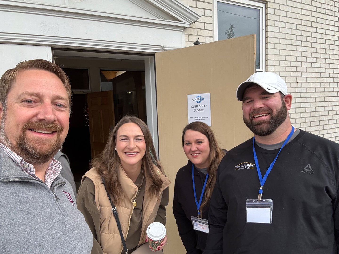 Four adults wearing name badges smile for a group selfie outside a building with a tan door. A “Keep Door Closed” sign is posted behind them. One woman holds a coffee cup. It appears to be cloudy outside.