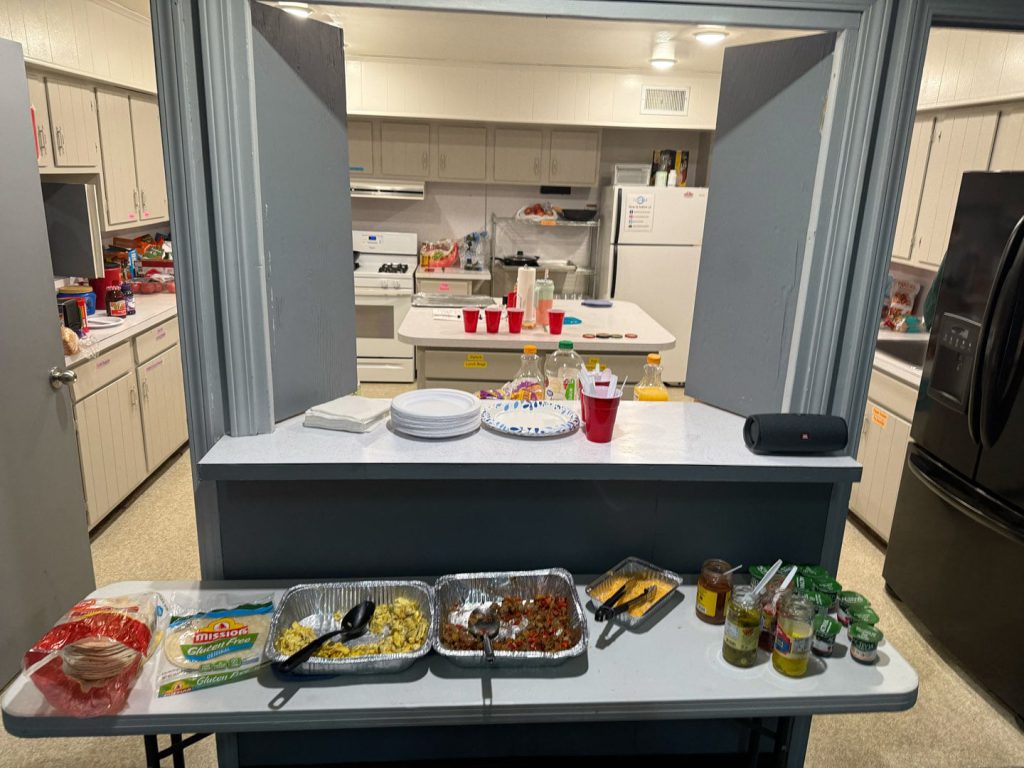 A kitchen with a serving counter displaying trays of food, tortillas, jars of condiments, and utensils. Behind the counter, the kitchen has white cabinets, a fridge, stove, and red cups on the counter.