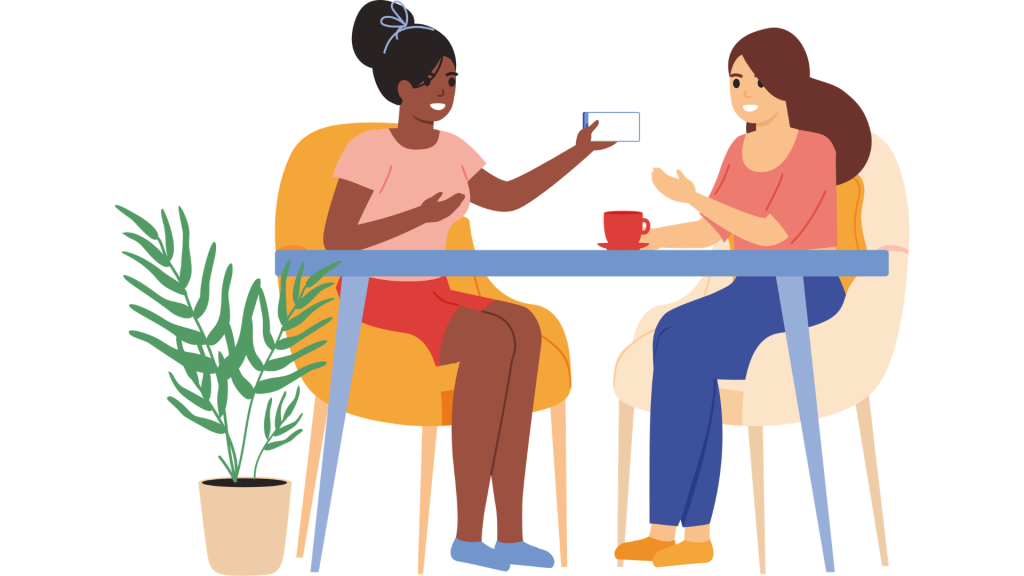 Two women sitting at a table having a conversation; one is holding up a smartphone, and the other has a red cup. A green potted plant sits beside them.