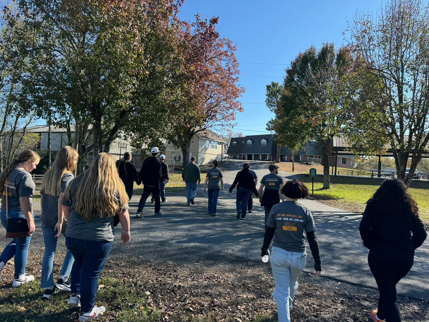 A group of people wearing matching gray T-shirts walk together on a paved path outside on a sunny day, surrounded by trees with autumn leaves.