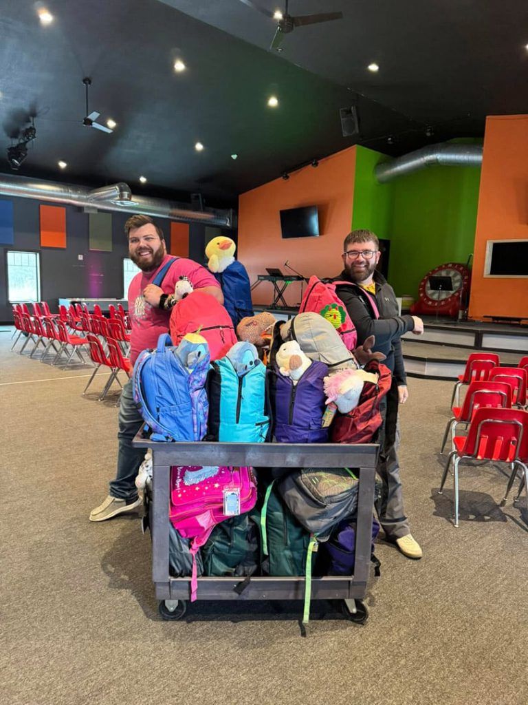 Two smiling men stand in a colorful room with rows of red chairs, posing behind a cart filled with rolled sleeping bags and backpacks. The setting appears to be a community center or event space.