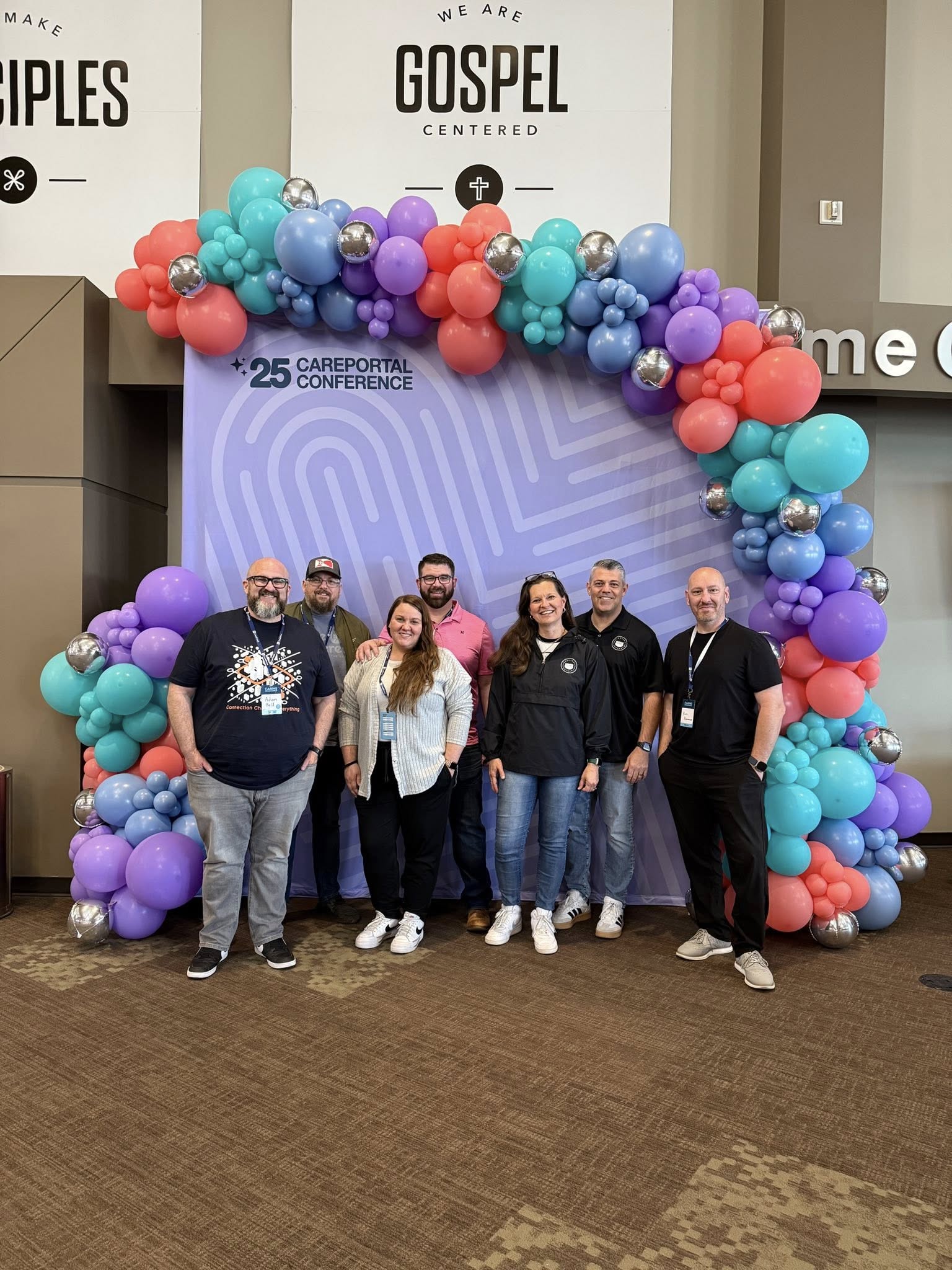 A group of seven people stand smiling in front of a purple CarePortal Conference backdrop decorated with colorful balloons. Large signs above read "We Are Disciples" and "We Are Gospel Centered.