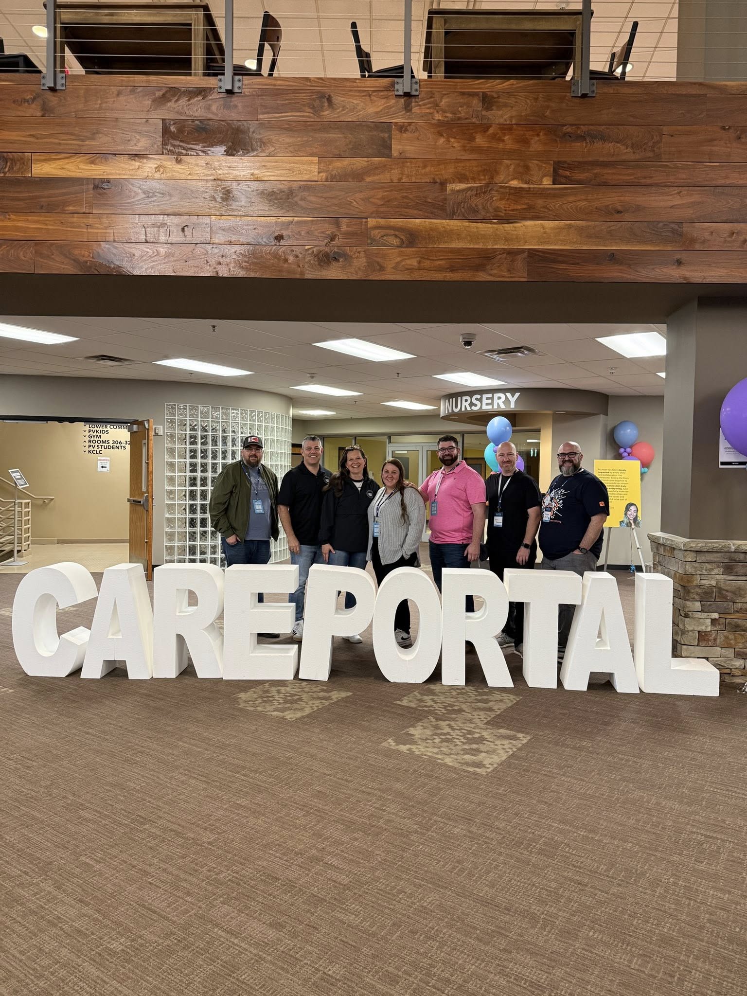 Seven people stand smiling behind large white letters spelling "CAREPORTAL" in a spacious indoor area with brown floors and wooden accents. Signs for a nursery and balloons are visible in the background.