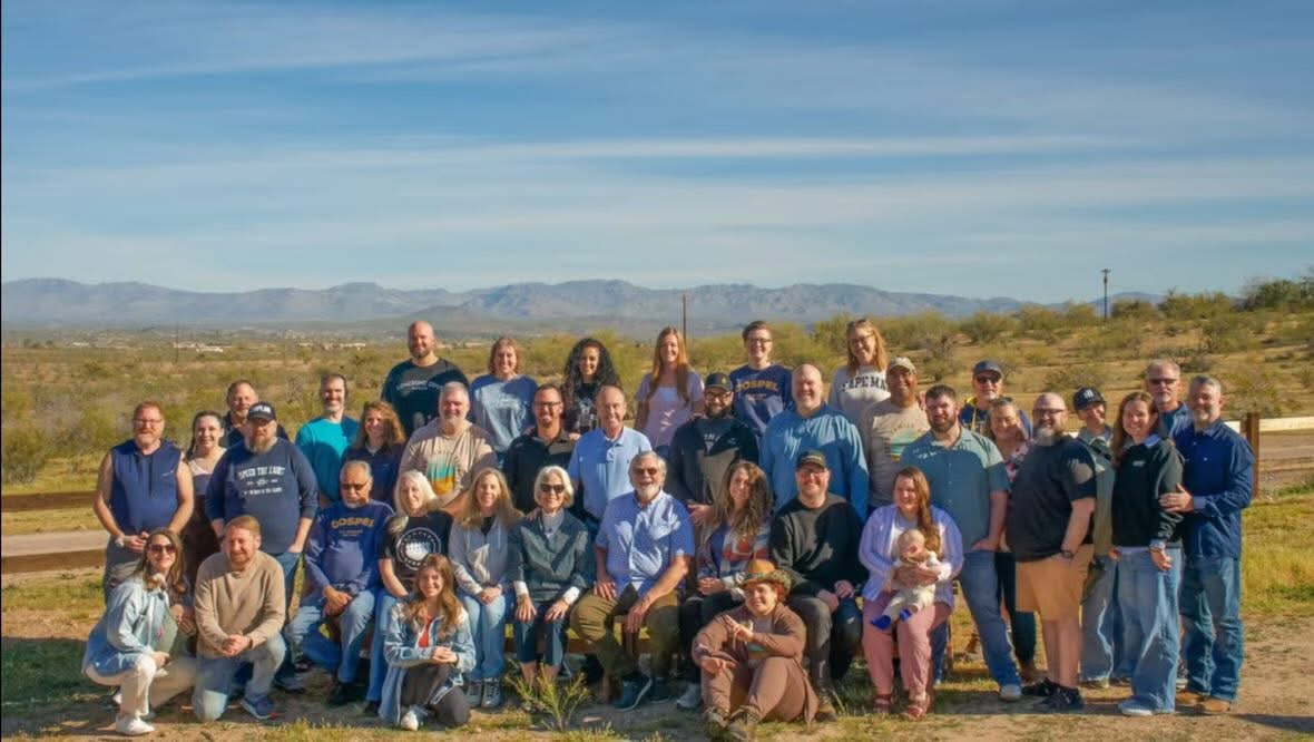 A large group of people of various ages pose outdoors on a sunny day, with mountains and a clear sky in the background. Some are seated on the grass while others stand behind them, all smiling for the photo.