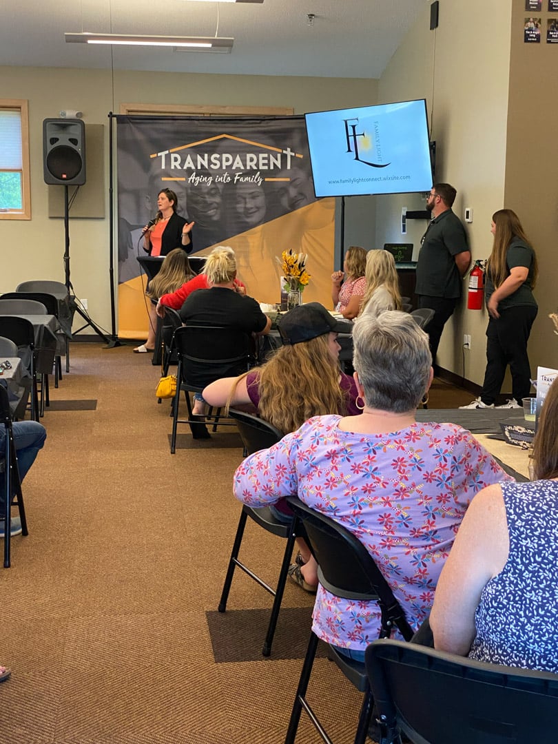 A woman stands and speaks to a seated audience in a meeting room. A banner behind her reads “TRANSPARENT Aging into Family.” Two people stand near a screen displaying a website, while attendees listen attentively.