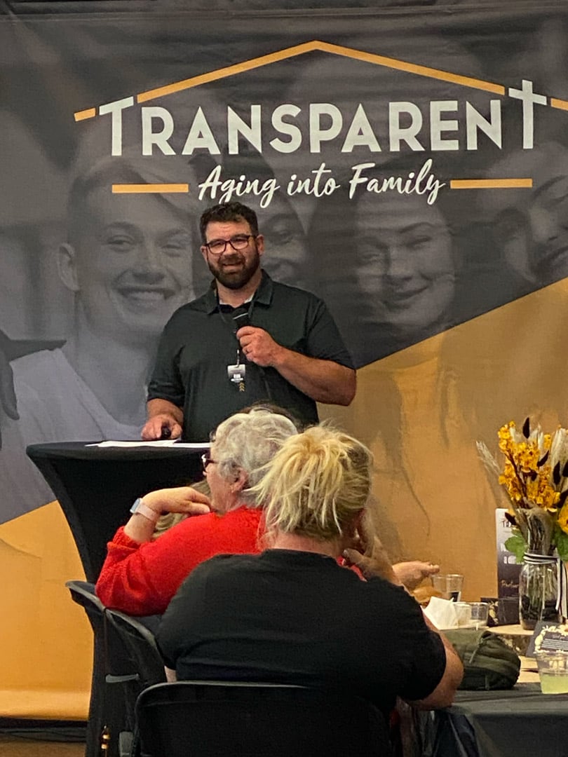 A man speaks at a podium in front of a banner that reads "TRANSPARENT Aging into Family," while people seated at tables listen. The setting appears to be a community or social event.
