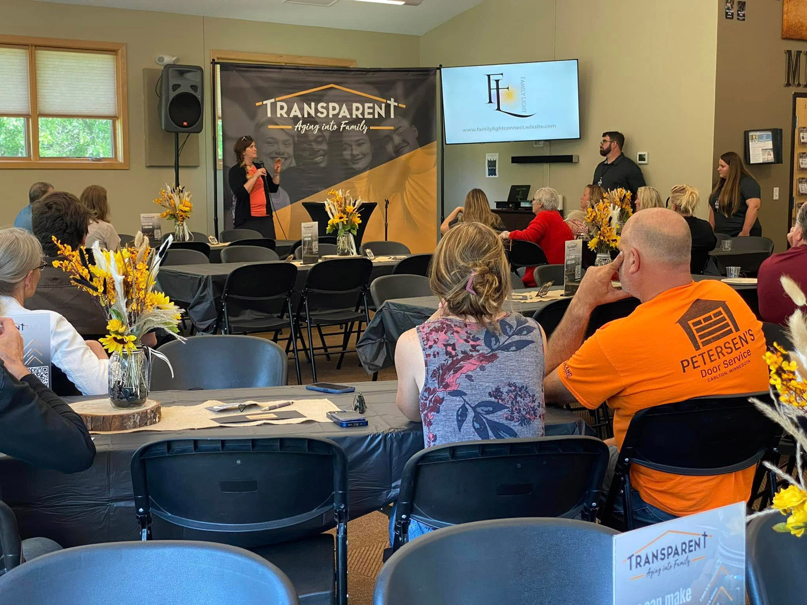 A woman speaks at the front of a room during a presentation, with people seated at tables decorated with yellow flowers. A large banner reads “TRANSPARENT” and a screen displays a logo and text.