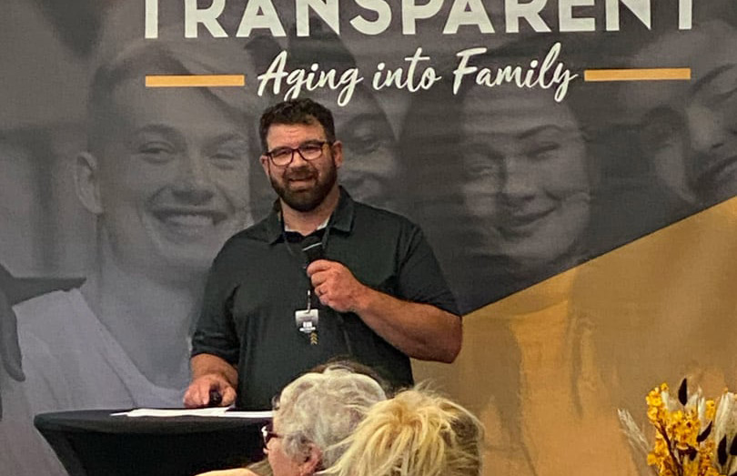 A man with glasses and a beard stands at a podium, speaking into a microphone in front of a banner that reads "TRANSPARENT Aging into Family." Several people are seated and listening in the foreground.