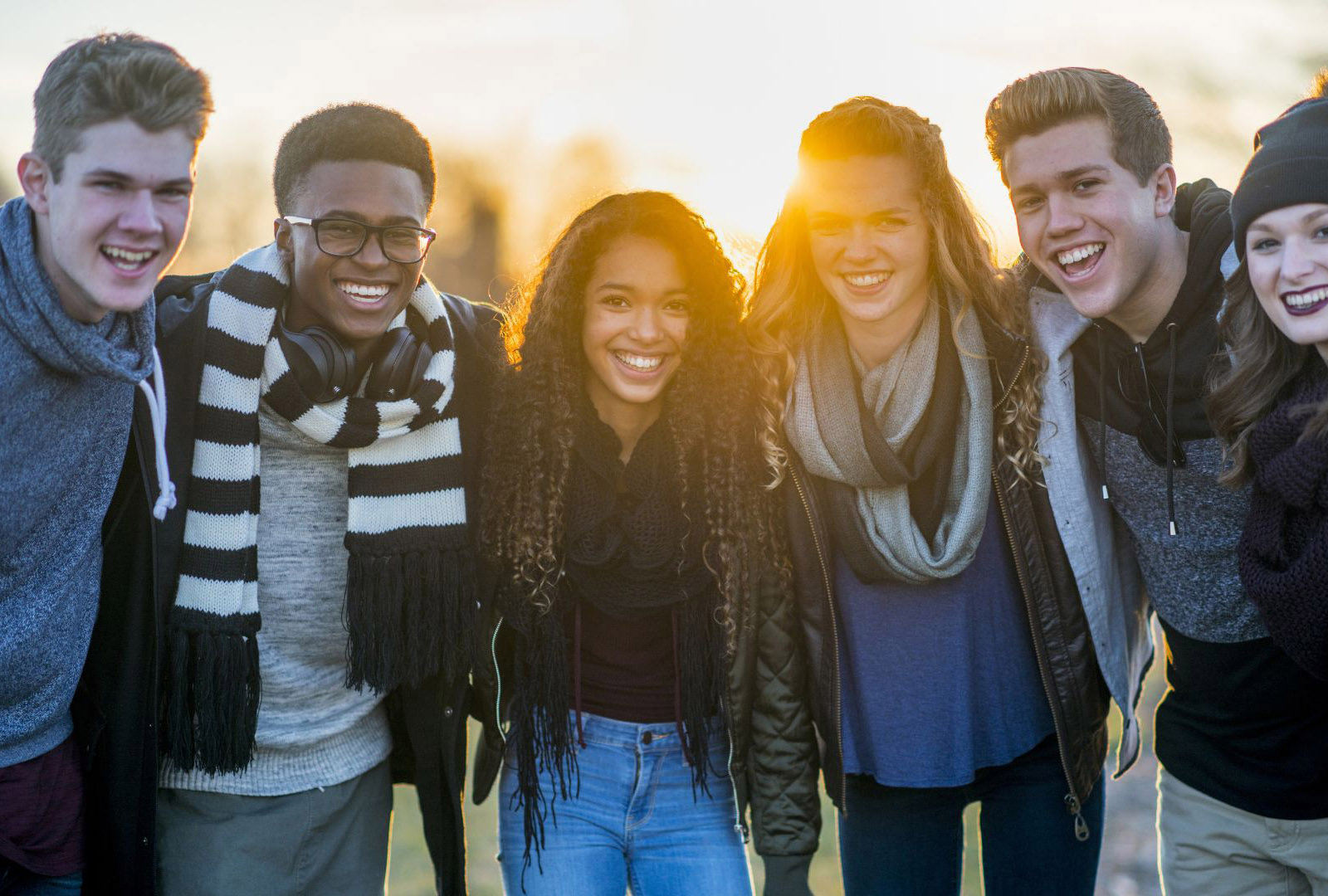 A group of six young adults stand close together outdoors, smiling and laughing, with the sun shining brightly behind them. They are dressed in casual winter clothes, including scarves and sweaters.