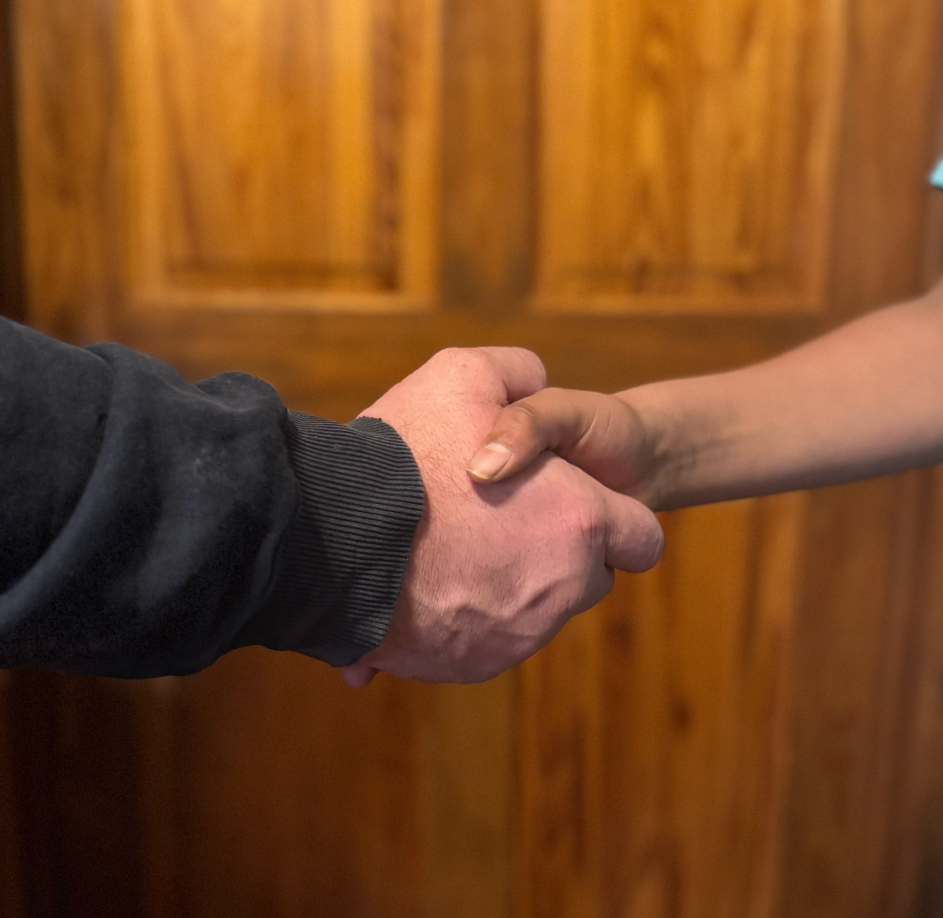 Two people shaking hands in front of a wooden door, one wearing a dark sleeve and the other with a bare arm.
