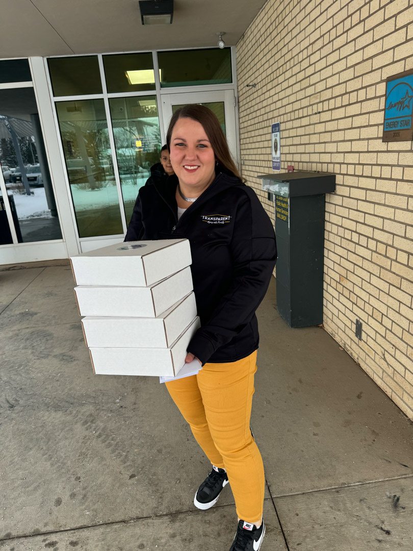 A woman in a black jacket and yellow pants smiles while holding a stack of four white boxes outside a building with a brick wall and glass doors. Snow is visible in the background.