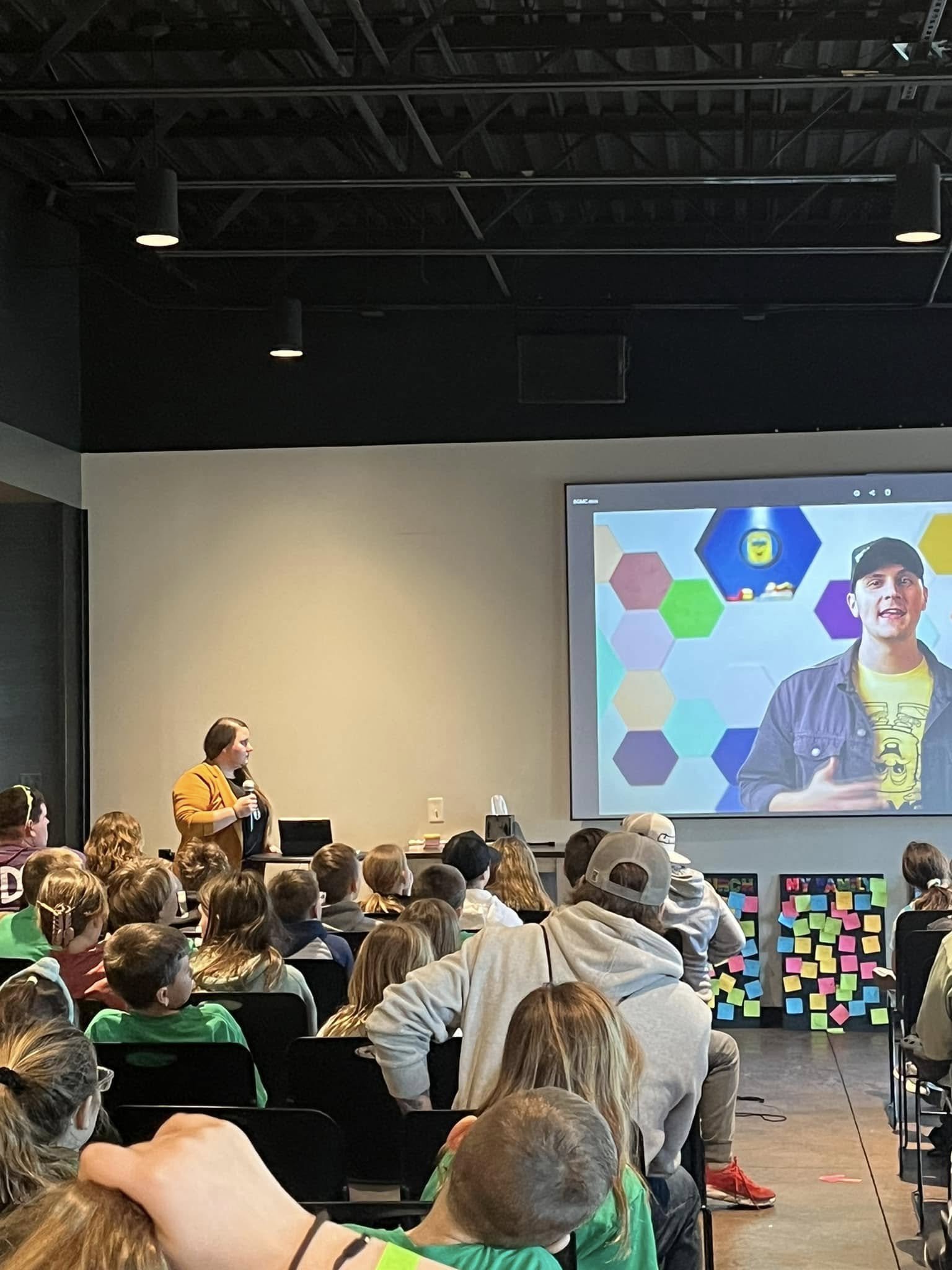 A woman speaks to a seated audience of children while a man appears on a large screen via video call. The screen displays colorful hexagons, and sticky notes are posted on the front wall below the screen.