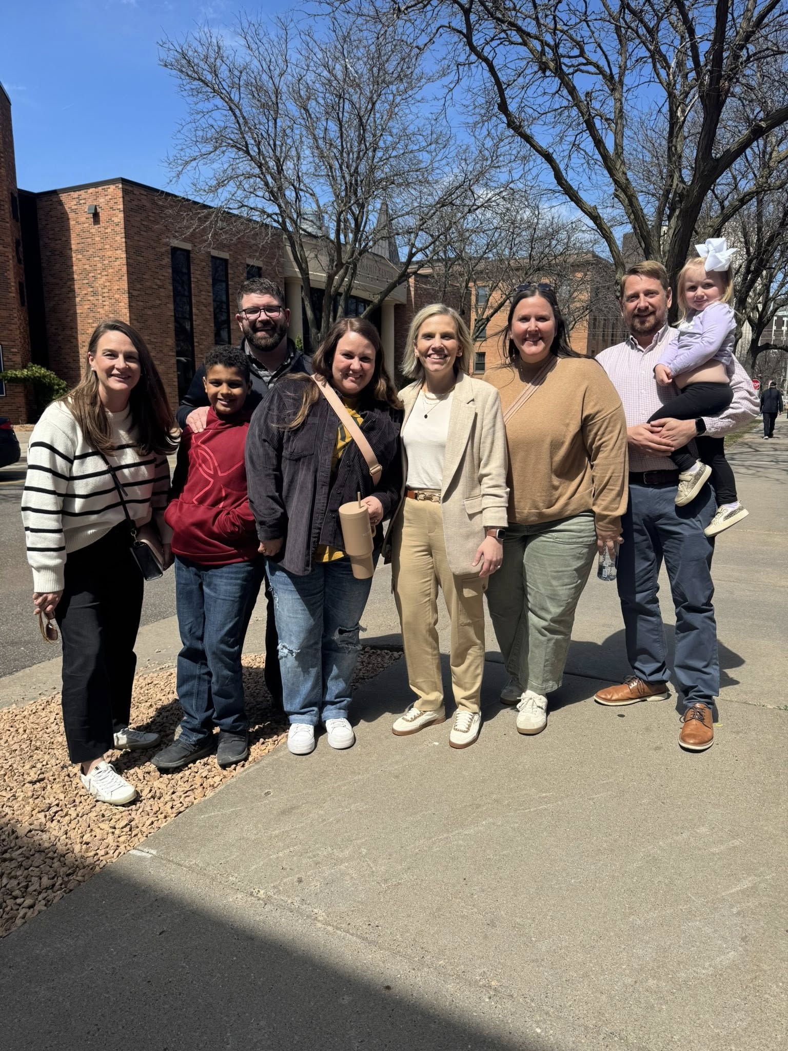 A group of seven adults and one child smiling and posing together outside on a sunny day, with buildings and leafless trees in the background.