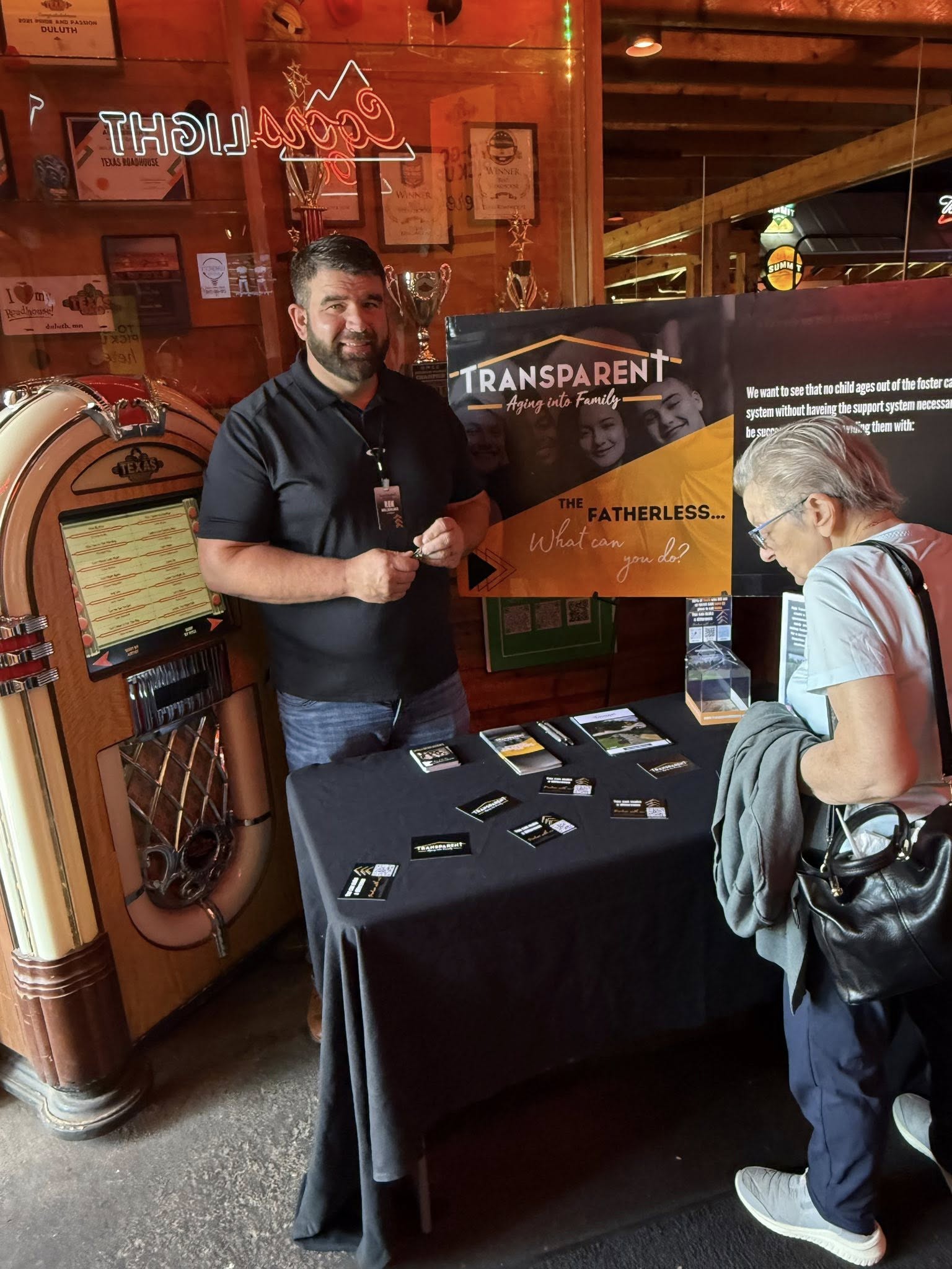 A man stands behind a display table with brochures and cards, talking to an older woman in a casual indoor setting. A large sign behind him reads “TRANSPARENT: The Fatherless… What can you do?” with a jukebox to the left.