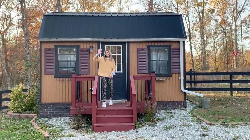 A woman stands on the front steps of a small brown and tan house with a black roof, waving and smiling. The house is surrounded by trees with autumn leaves and a fenced yard.