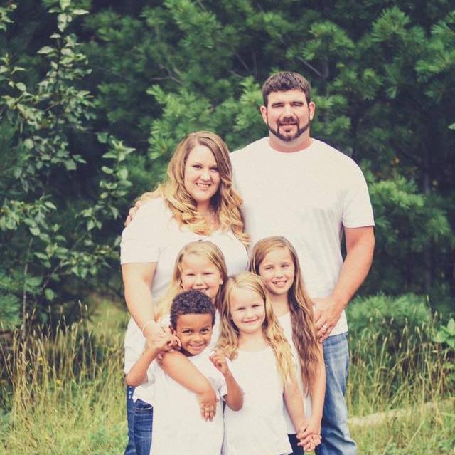 A smiling family of two adults and four children, all wearing white shirts, poses together outdoors in front of green trees and grass.