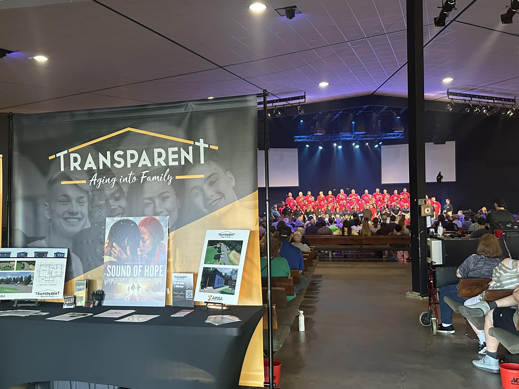 A booth with a "TRANSPARENT Aging into Family" banner stands in the foreground of a large indoor venue. In the background, a choir in red attire performs on stage for an audience seated on benches.