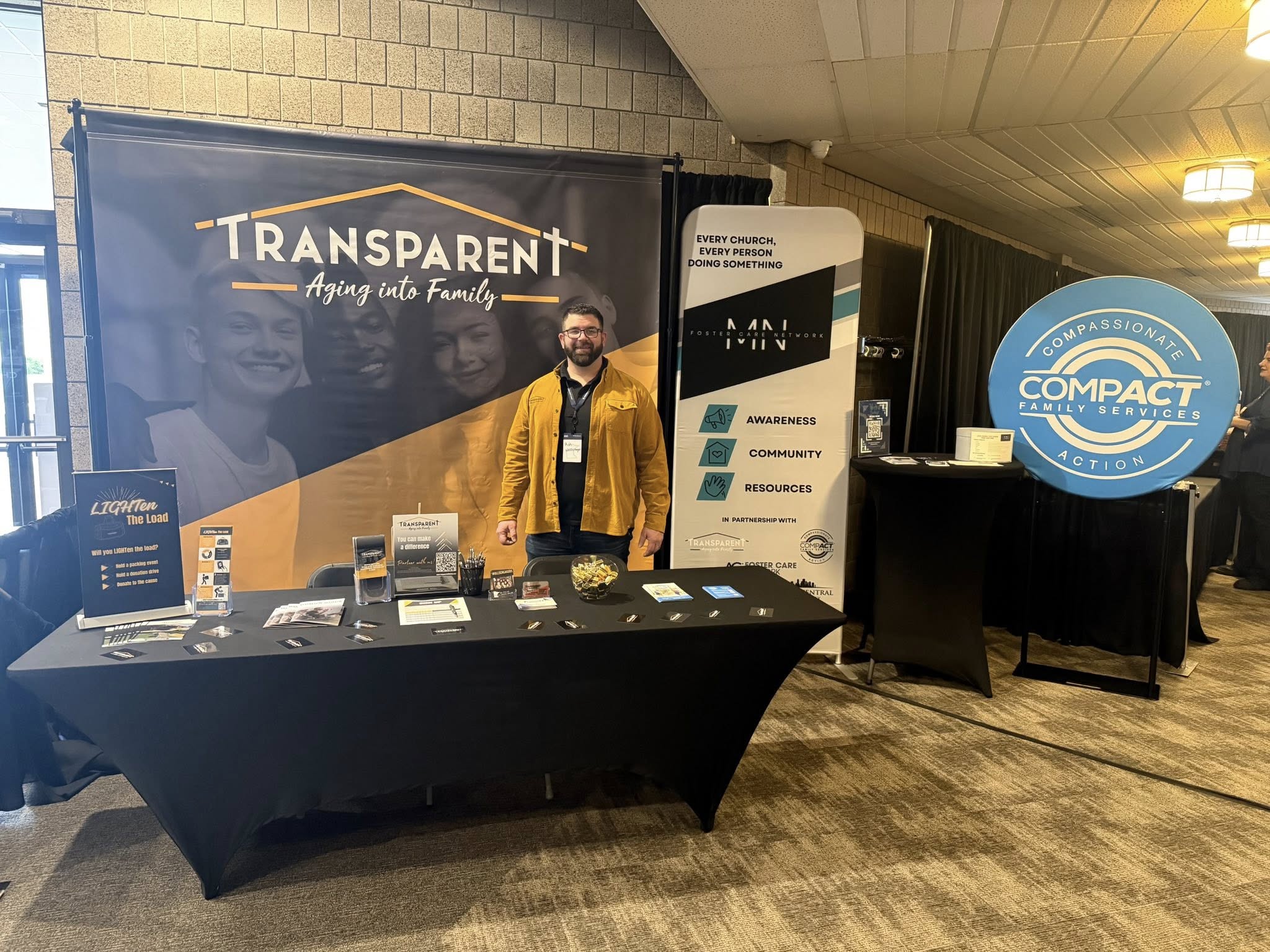 A man stands and smiles behind a display table for "Transparent: Aging into Family" at an indoor event. The table holds brochures and candy. Two banners for related organizations are visible nearby.