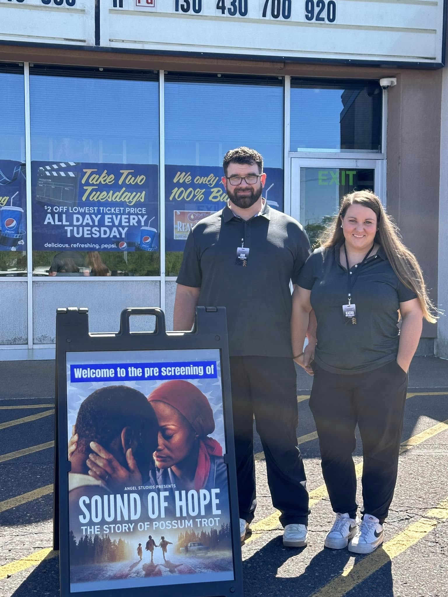 Two people in matching uniforms stand outside a building next to a sign promoting the movie "Sound of Hope: The Story of Possum Trot." The building has large windows and a marquee above.