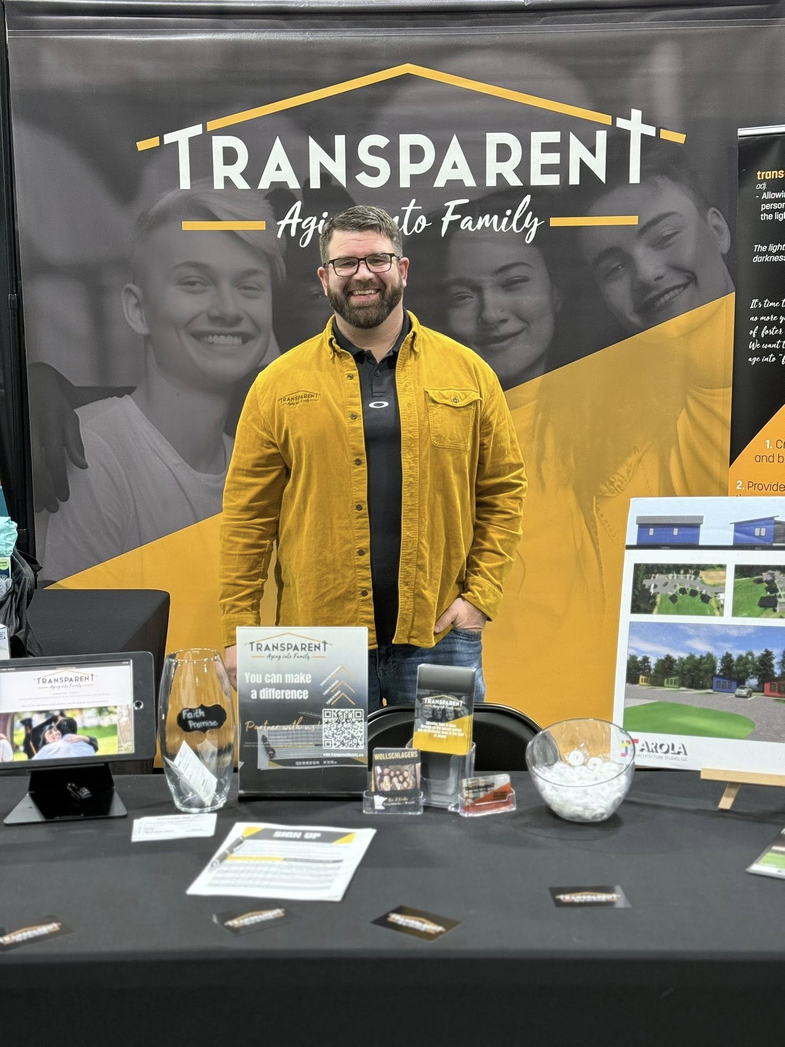 A man in a yellow jacket stands smiling at a booth for "Transparent Aging Into Family," with brochures, flyers, a jar, and display boards on the table in front of him. A large branded banner is in the background.