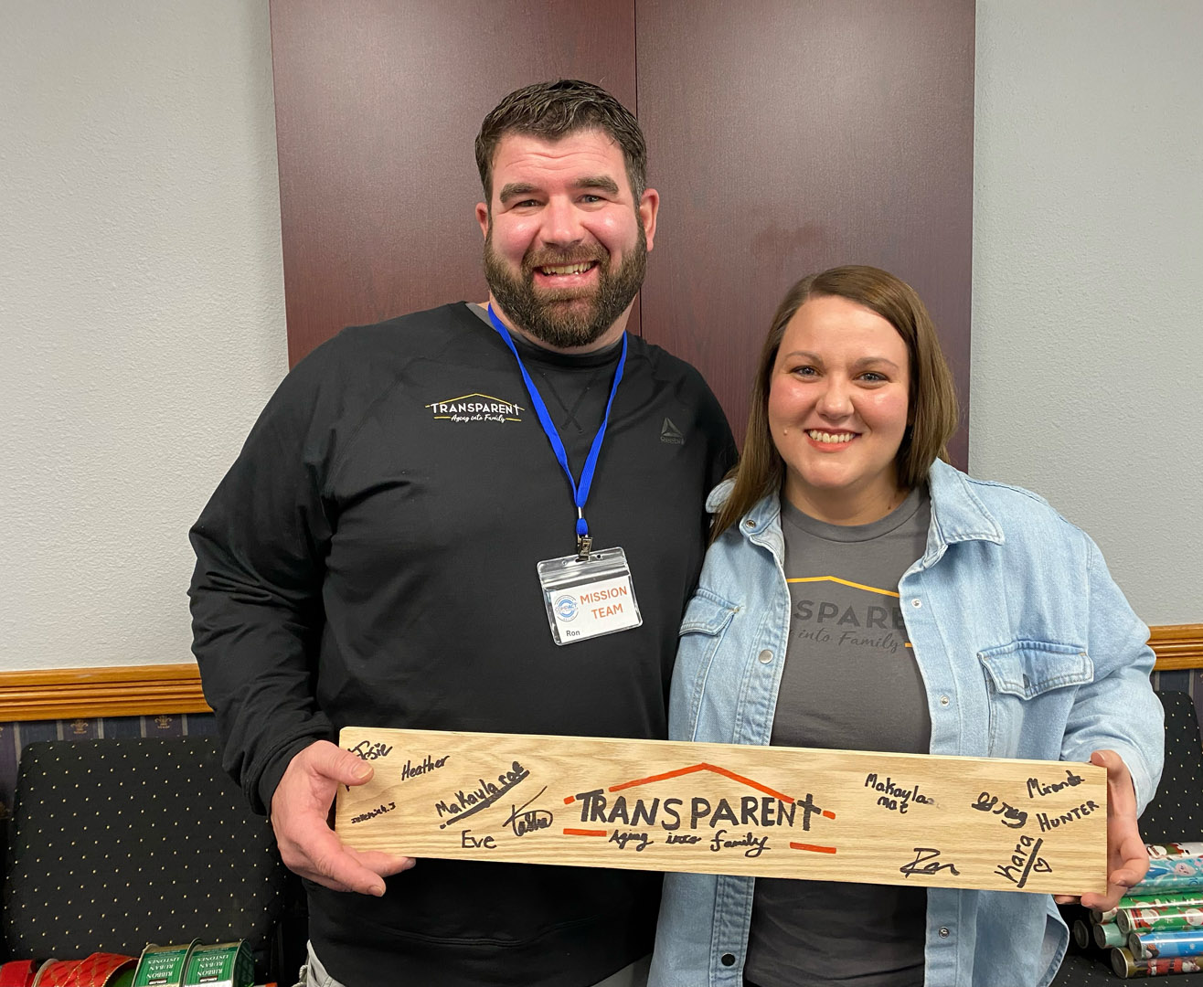 Two smiling adults stand indoors holding a wooden sign that reads "TRANSPARENT: Hope. Love. Family." The sign is decorated with various handwritten names. Both wear casual clothing and the man has a mission team badge.