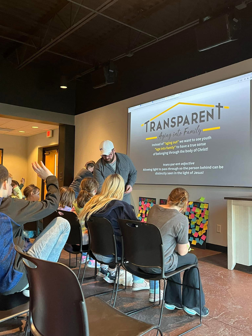 A group of young people sit and raise their hands in a classroom, facing a projector screen displaying a presentation titled "TRANSPARENT: Aging into Family" with a man standing at the front, engaging with the students.