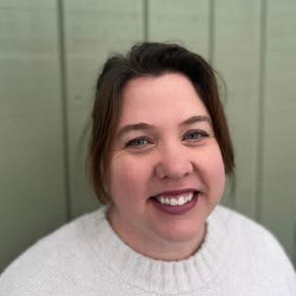 A woman with shoulder-length brown hair smiles at the camera. She is wearing a white sweater and standing in front of a light green paneled wall.