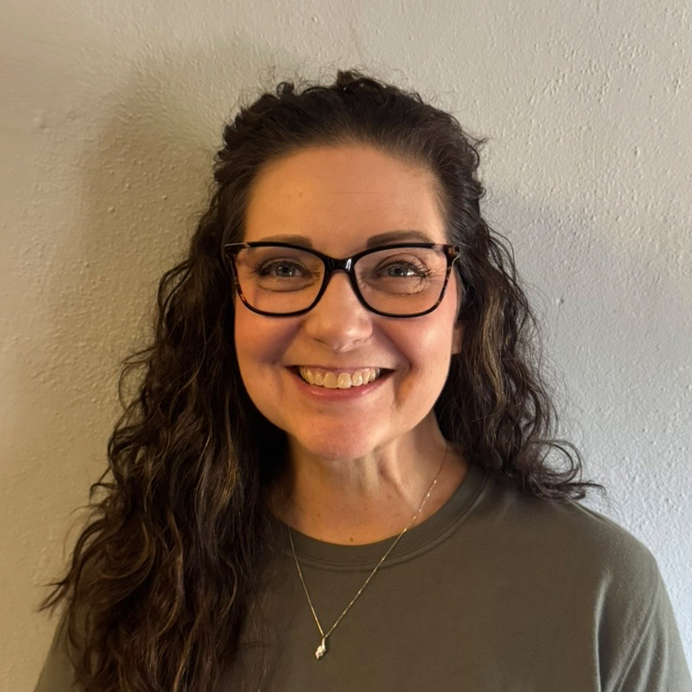 A woman with long curly brown hair and glasses smiles at the camera. She is wearing a gray top and a necklace with a small pendant, standing against a light-colored wall.