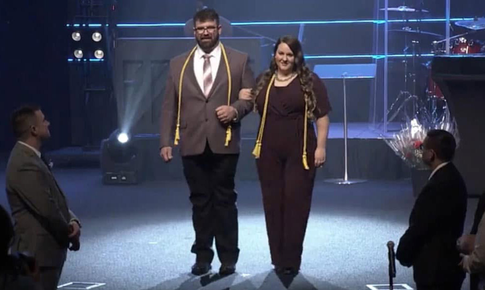 Two people, a man and a woman dressed in formal attire with honor cords, walk arm-in-arm down a stage aisle as an audience looks on. Stage lighting and musical instruments are visible in the background.