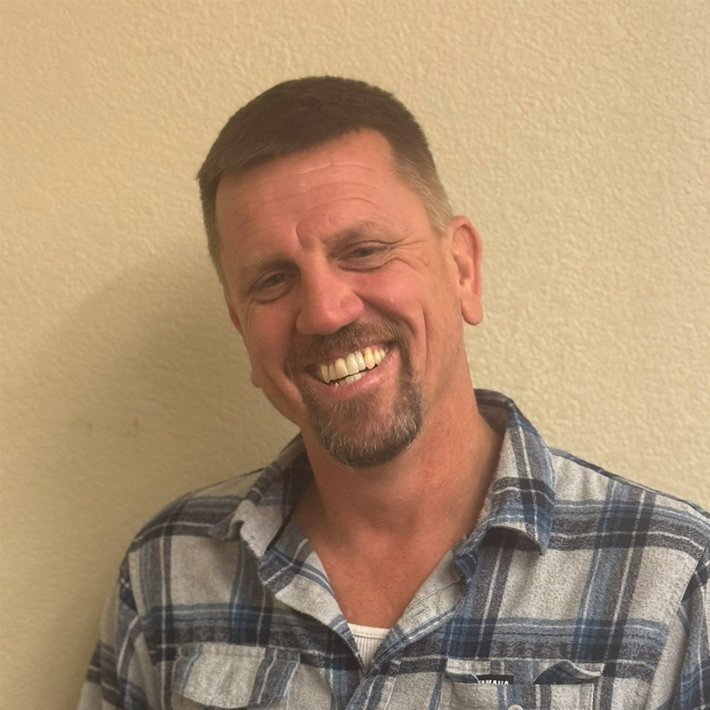 A middle-aged man with short hair and a goatee smiles at the camera, wearing a blue and gray plaid shirt against a plain beige wall.