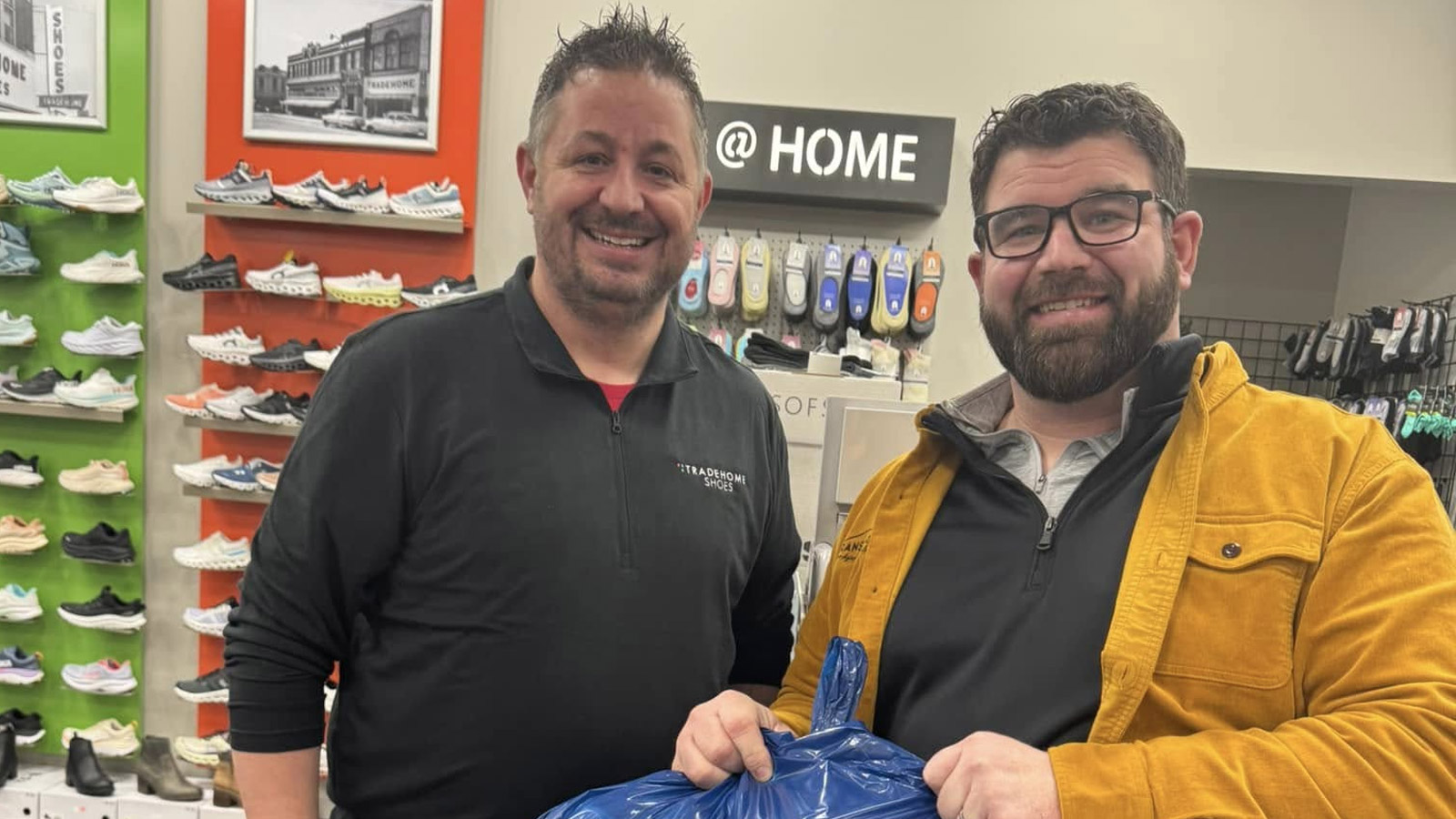 Two smiling men stand in a shoe store, one holding a blue bag. Shelves with various shoes are visible in the background, along with a sign that says "@ HOME" above them.