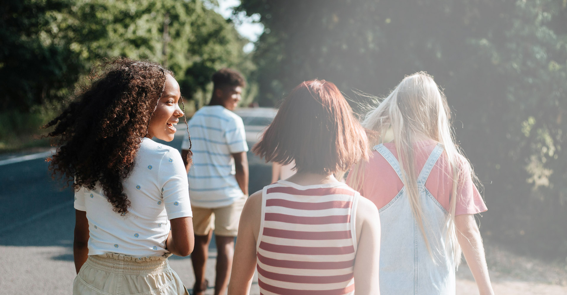 Four young people walk along a sunlit road surrounded by greenery. Two walk ahead, while two, including one smiling, walk behind them. They all wear casual summer clothes. The scene feels relaxed and cheerful.