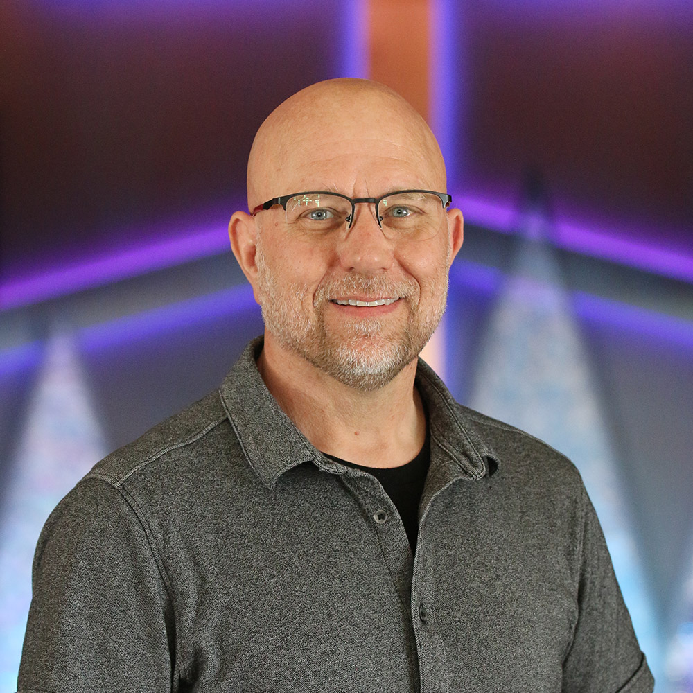 A middle-aged man with glasses, a bald head, and a short beard smiles at the camera. He is wearing a dark gray collared shirt. The background features blurred purple and blue lights in geometric patterns.
